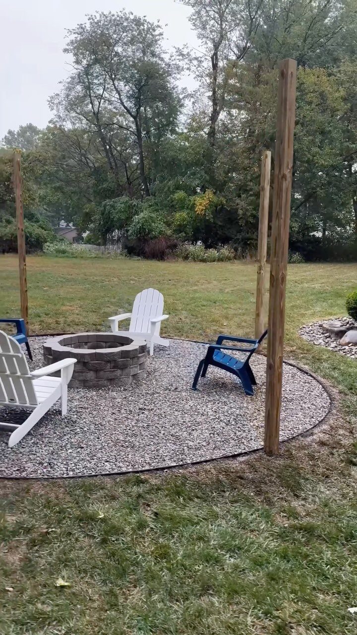 A stone fire pit surrounded by chairs on a gravel patio in a backyard.