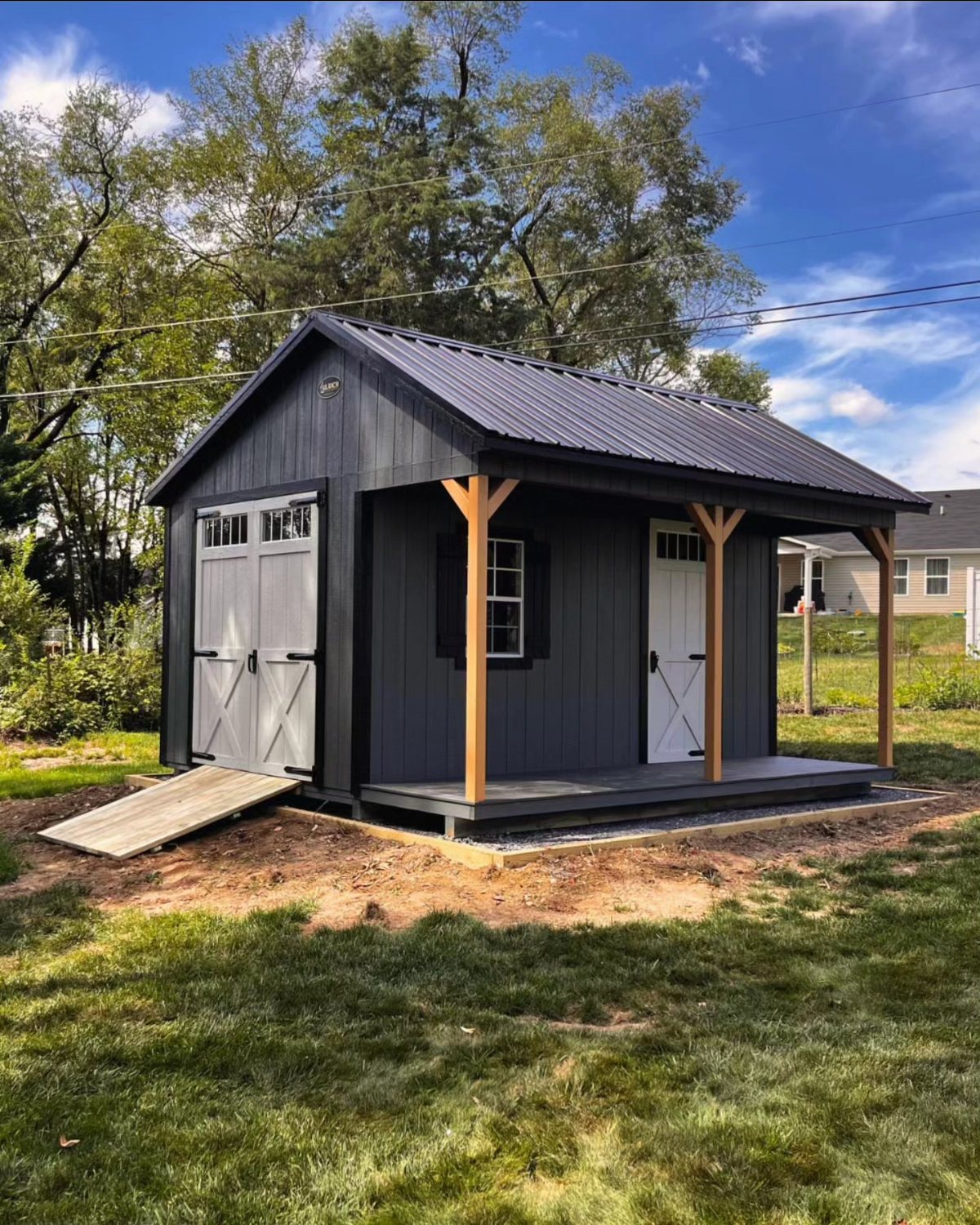 A dark grey wooden backyard shed featuring a small porch with light wood support posts and a ramp.