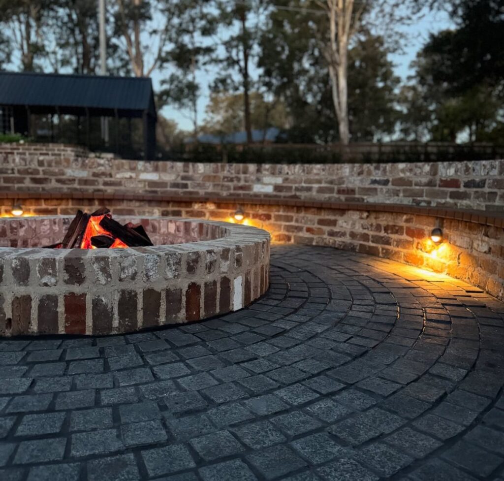 A circular brick fire pit with burning wood on a dark cobblestone patio at dusk.