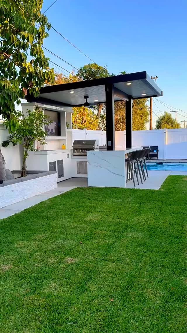 Modern outdoor kitchen with a built-in grill, marble countertops, and bar stools next to a pool.