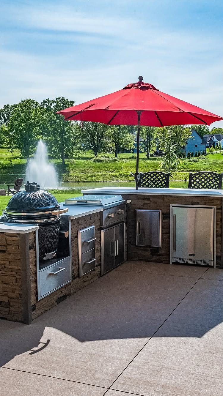 Built-in outdoor kitchen with stone cabinetry, stainless steel appliances, and a red patio umbrella on a concrete patio.