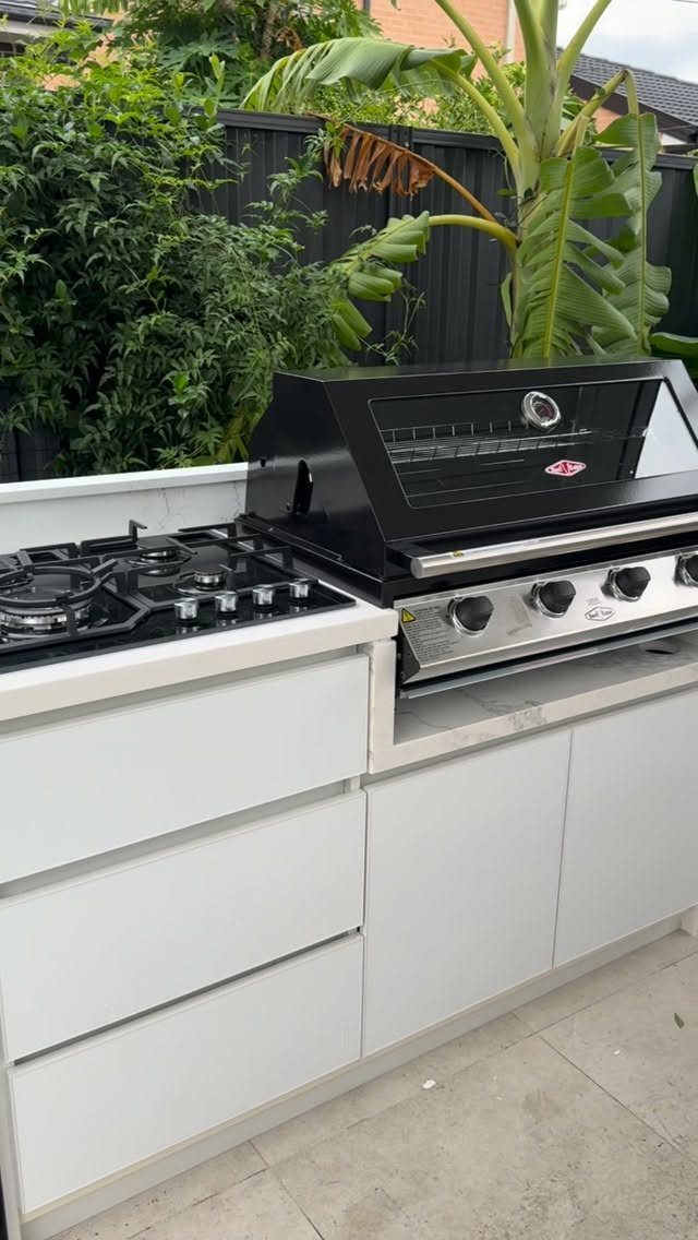 A compact outdoor kitchen with a white countertop, built-in grill, and gas cooktop near tropical plants.