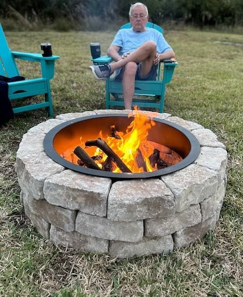 A stone fire pit with a burning fire sits on a grassy lawn near a man.