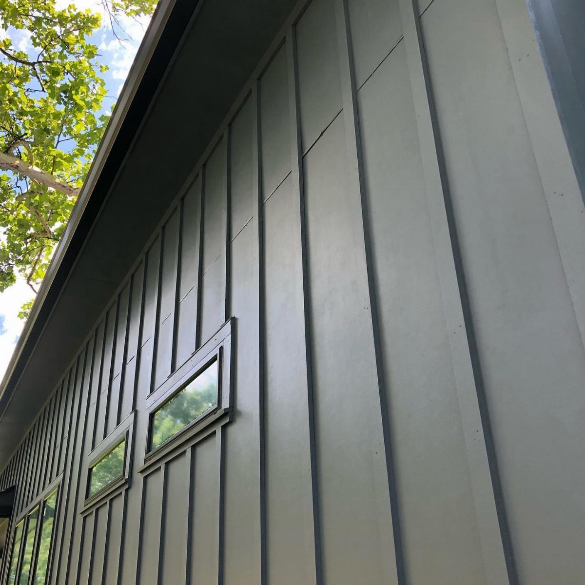 Dark gray board and batten siding on a building exterior with small windows and green trees.
