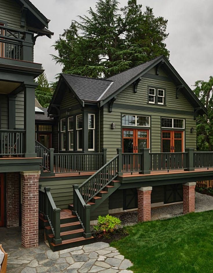 A dark green house with horizontal siding, a large wooden deck, and dark trim surrounded by trees.