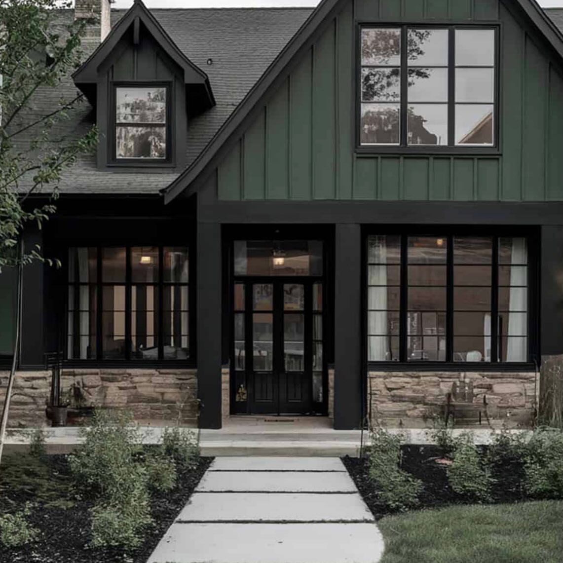 Dark green house with black window frames, stone base, and a concrete walkway leading to the front door.