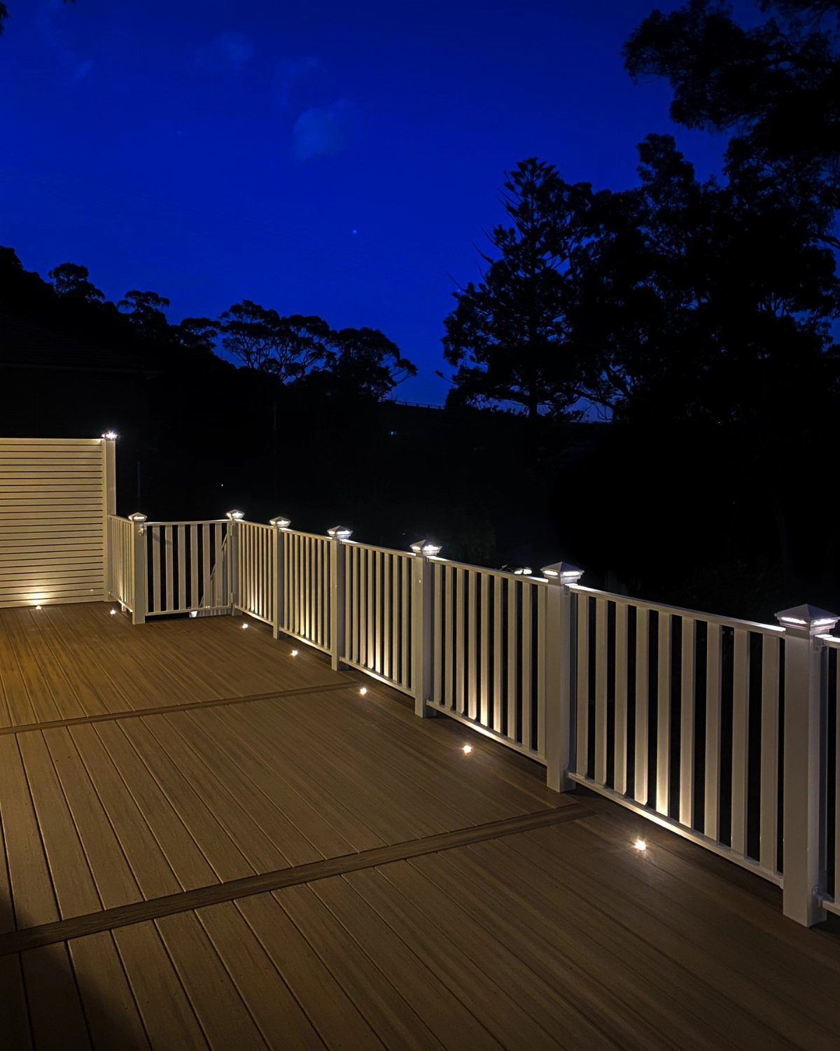 Wooden deck with white railings featuring integrated floor and post lighting at twilight.