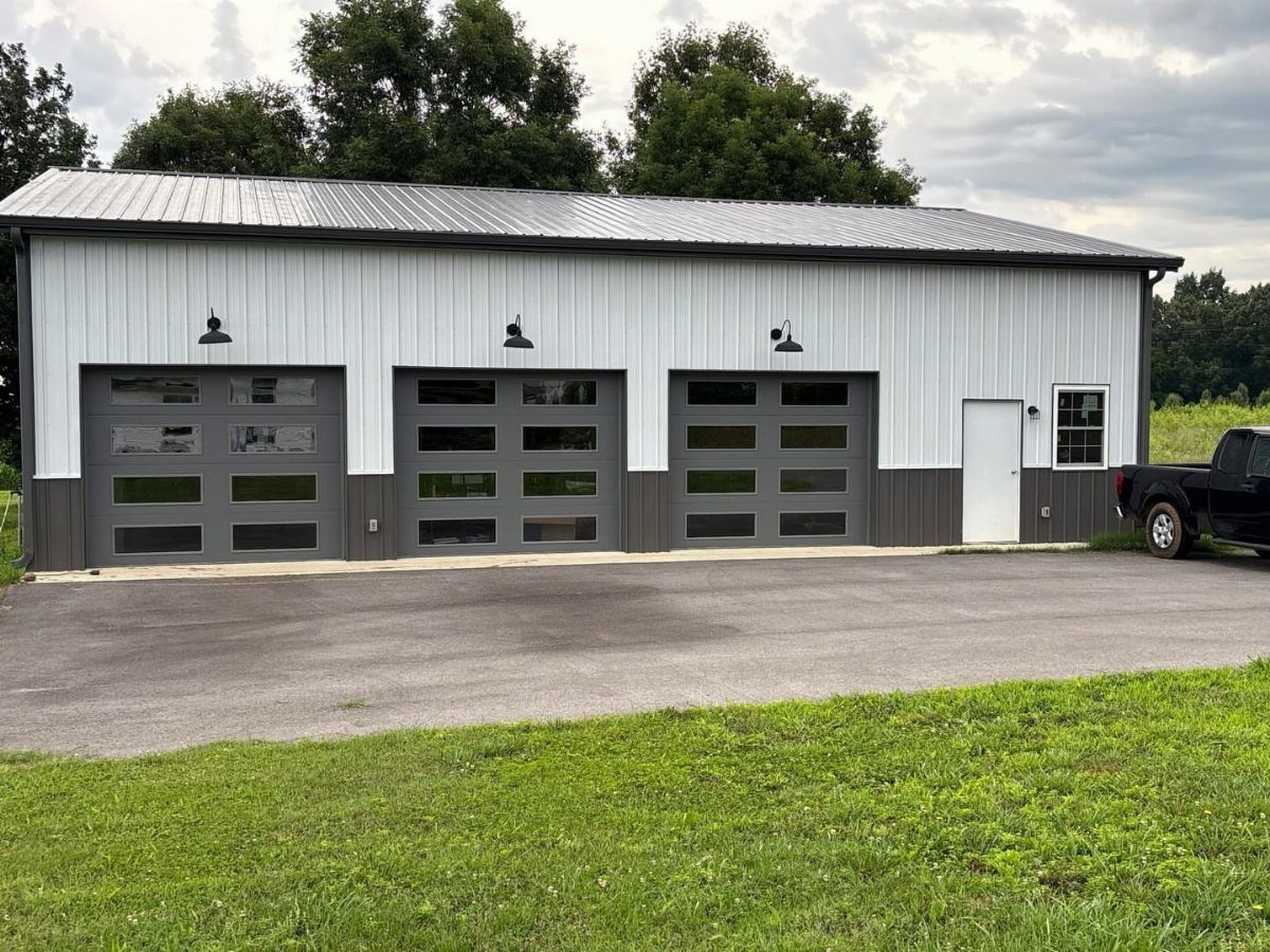 A white pole barn garage featuring three modern glass-paneled doors and black exterior lighting.