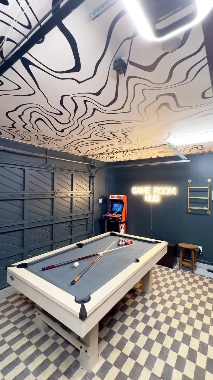 A blue pool table sits in a garage game room with an arcade machine and glowing sign.