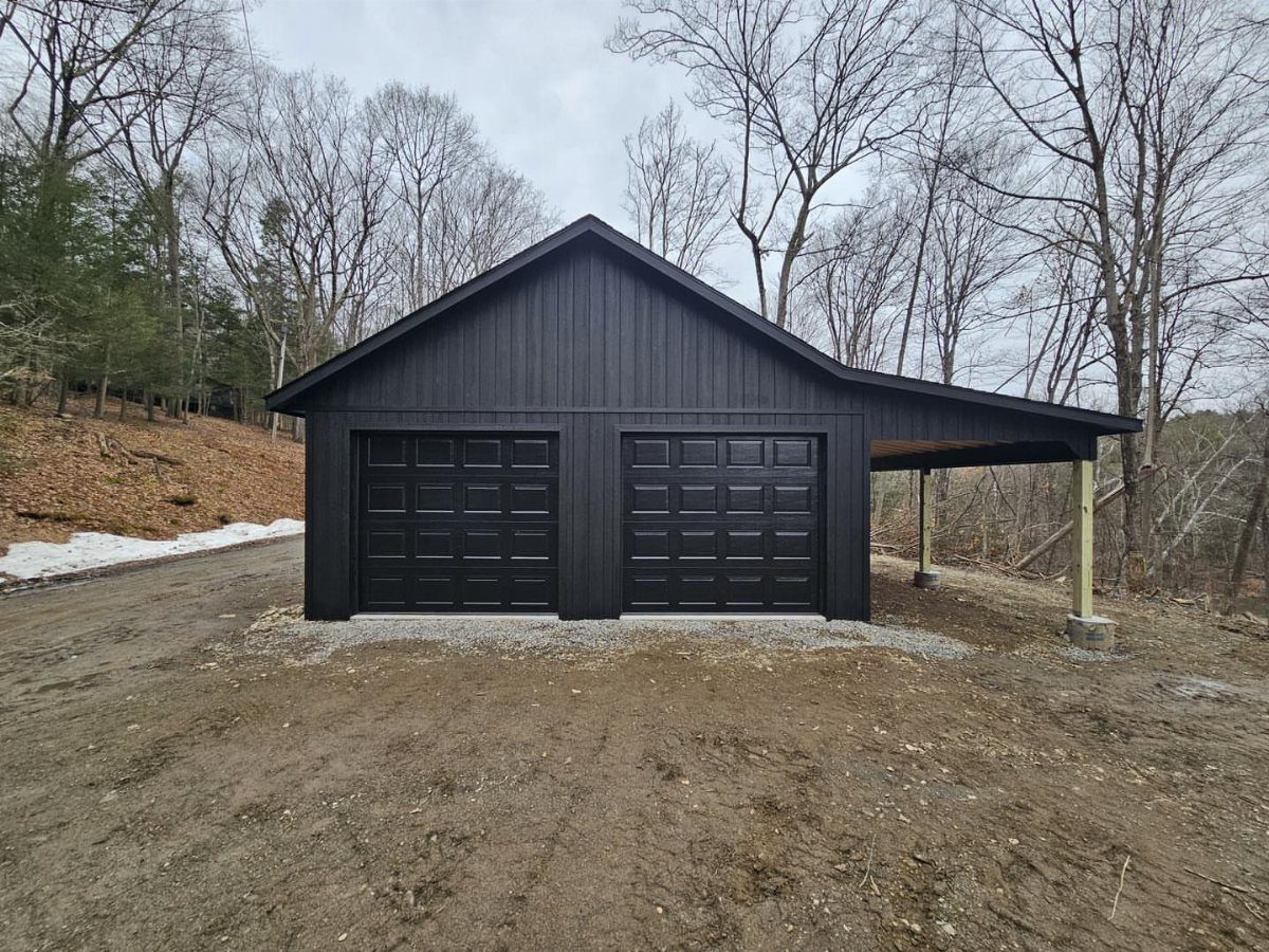 Black barn style garage with two doors and an attached lean-to structure under bare trees.