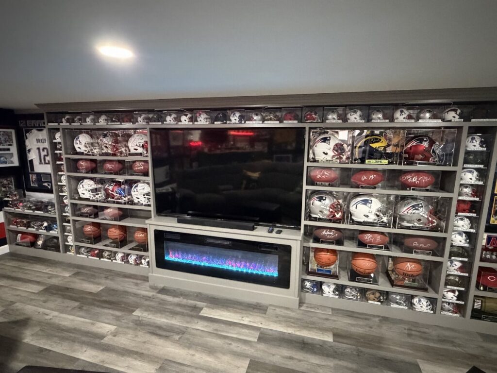 Grey shelves displaying various football helmets and footballs surrounding a large television and electric fireplace.
