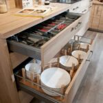 Open light-wood kitchen drawers with grey organizers holding plates, bowls, and utensils.