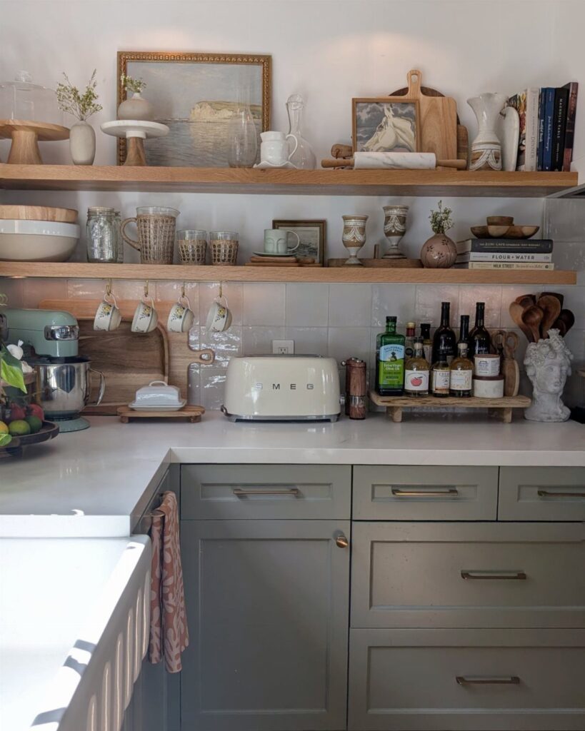 Natural wood open shelves holding dishes and decor above sage green kitchen cabinets.