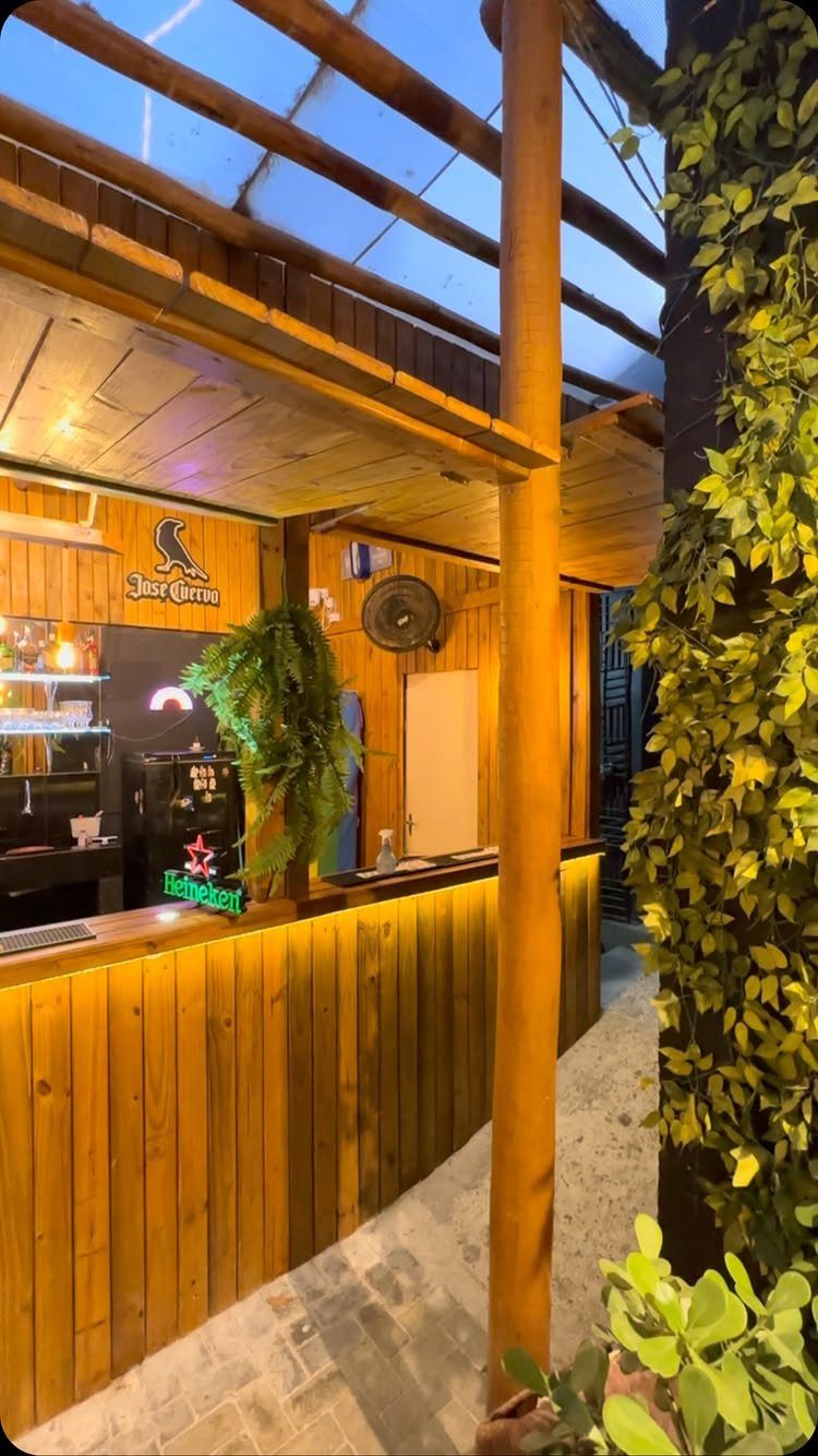 A wooden outdoor bar counter with warm lighting and green plants under a wooden structure.