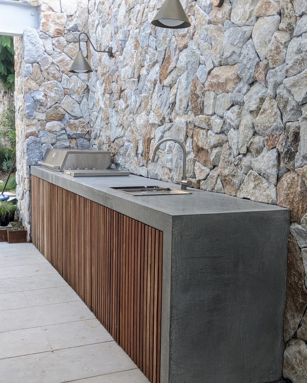 Grey concrete countertop with an integrated sink and wooden slat base against a stone wall.