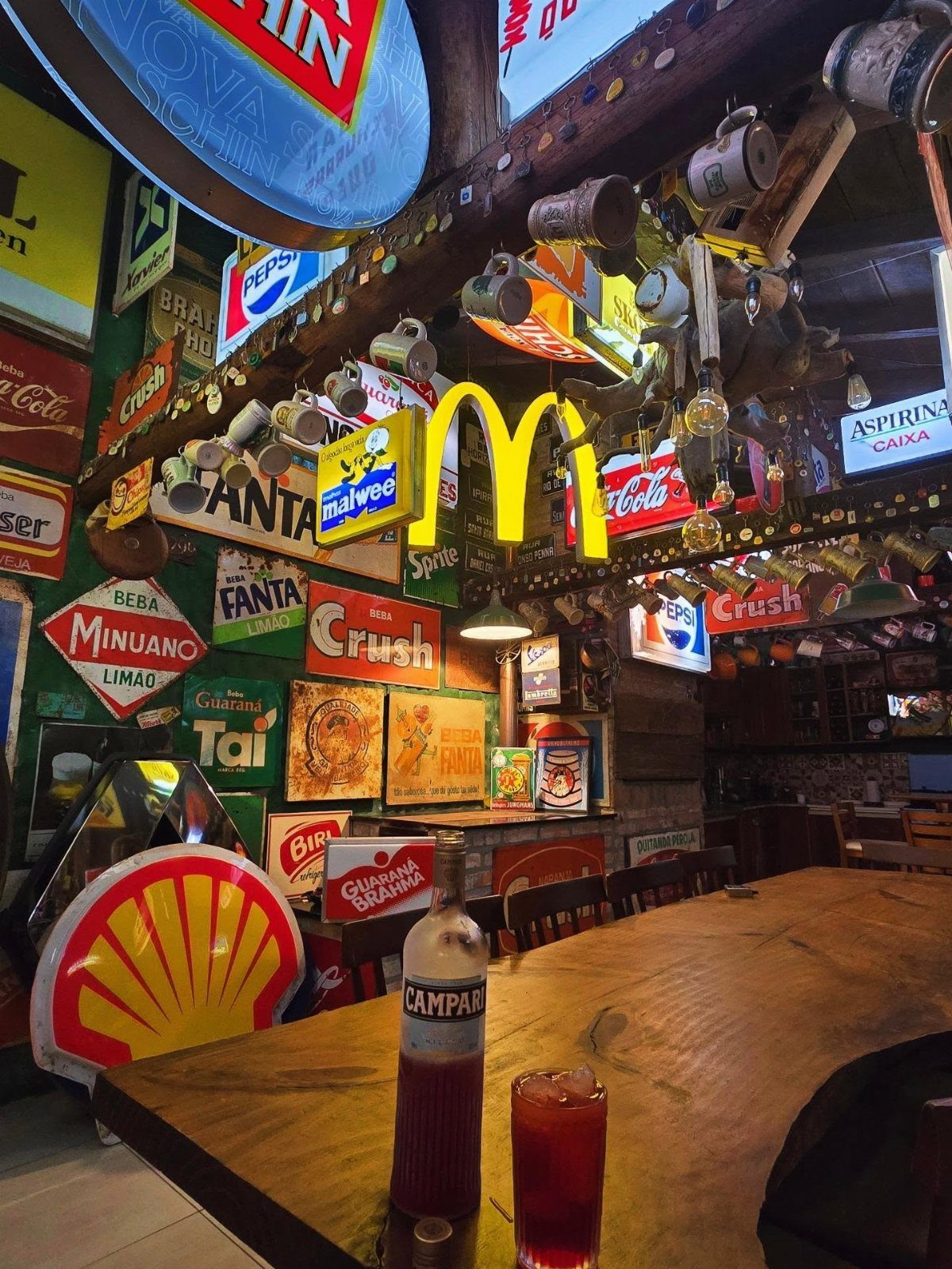 Vintage brand signs and advertisements decorate a bar area with a wooden counter and red cocktail.