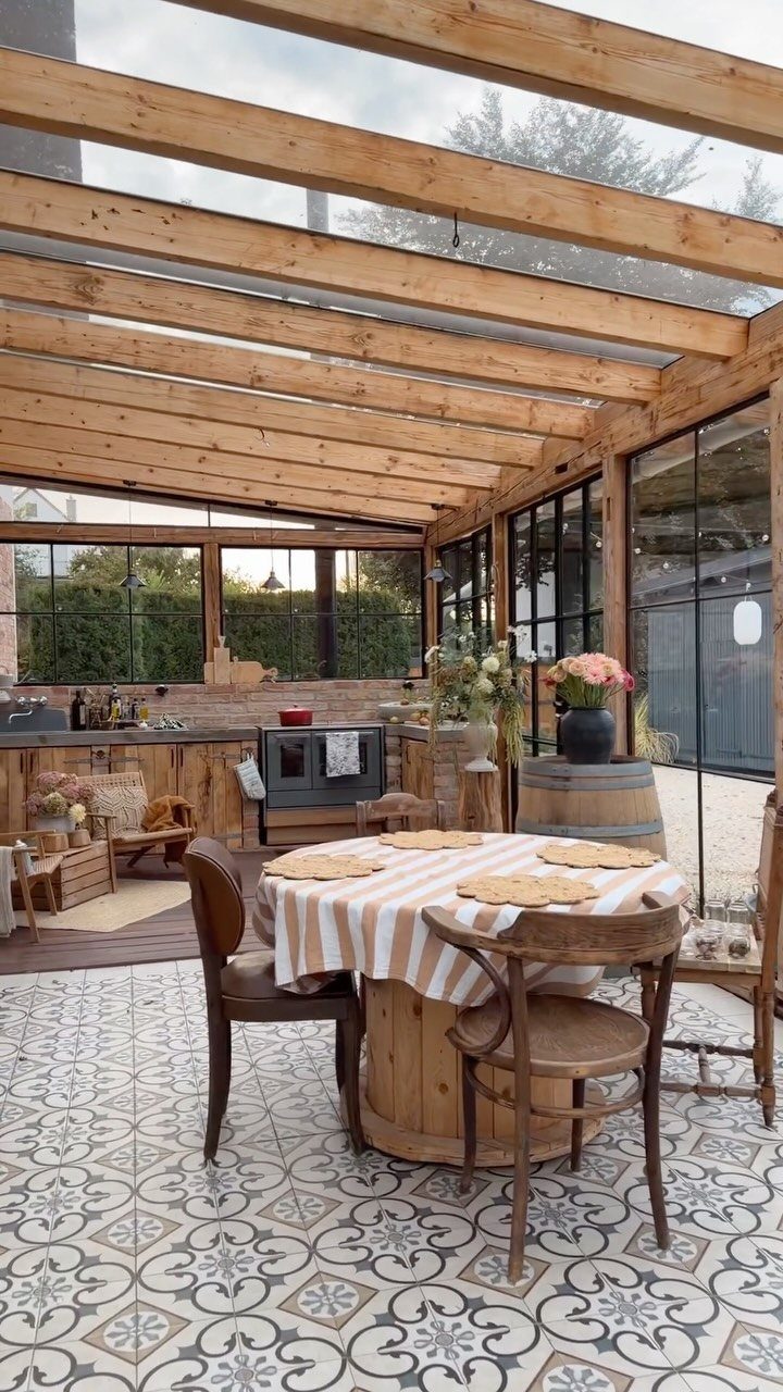 Rustic outdoor kitchen with wooden pergola, patterned tiles, and a wooden dining set under a glass roof.