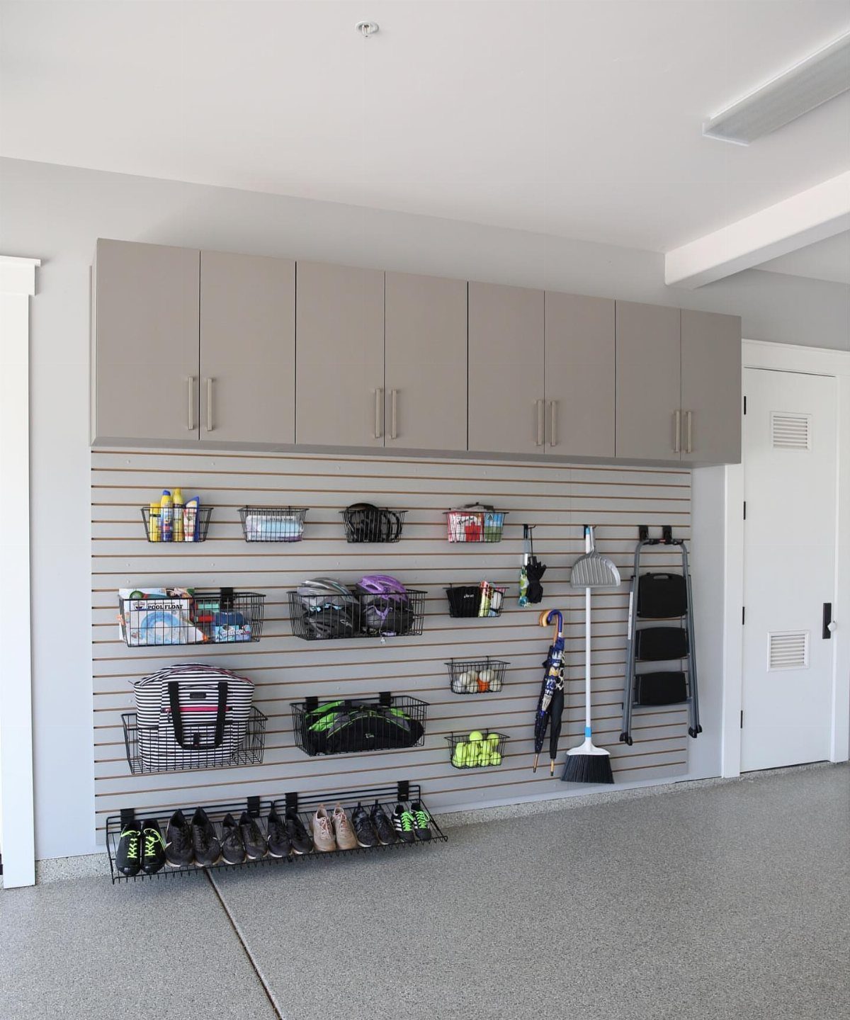 Grey slatwall storage system with hanging baskets, hooks, and tools mounted below grey cabinets in a garage.
