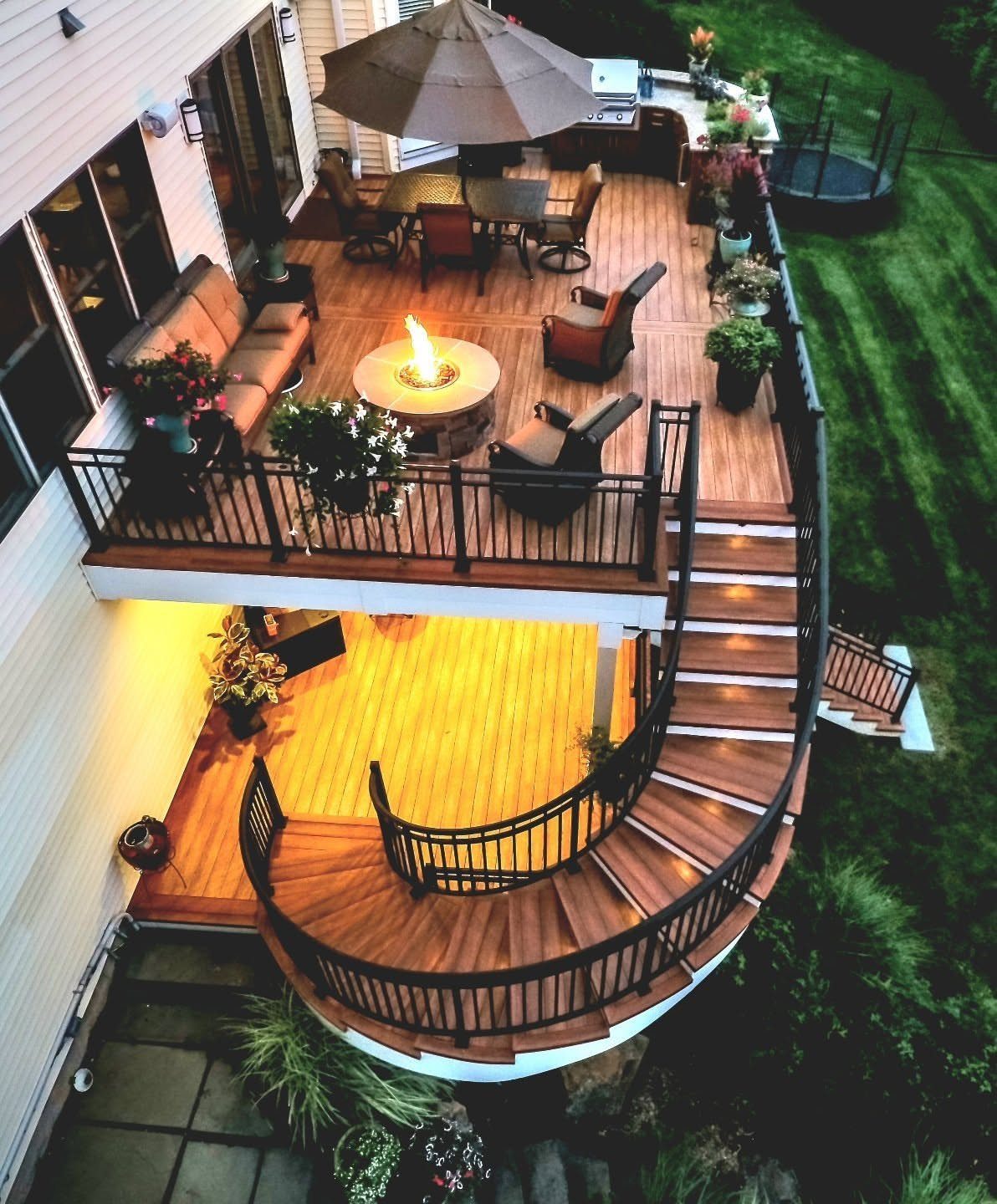 Two-level wooden deck with a fire pit, dining area, and curved stairs overlooking a green yard.