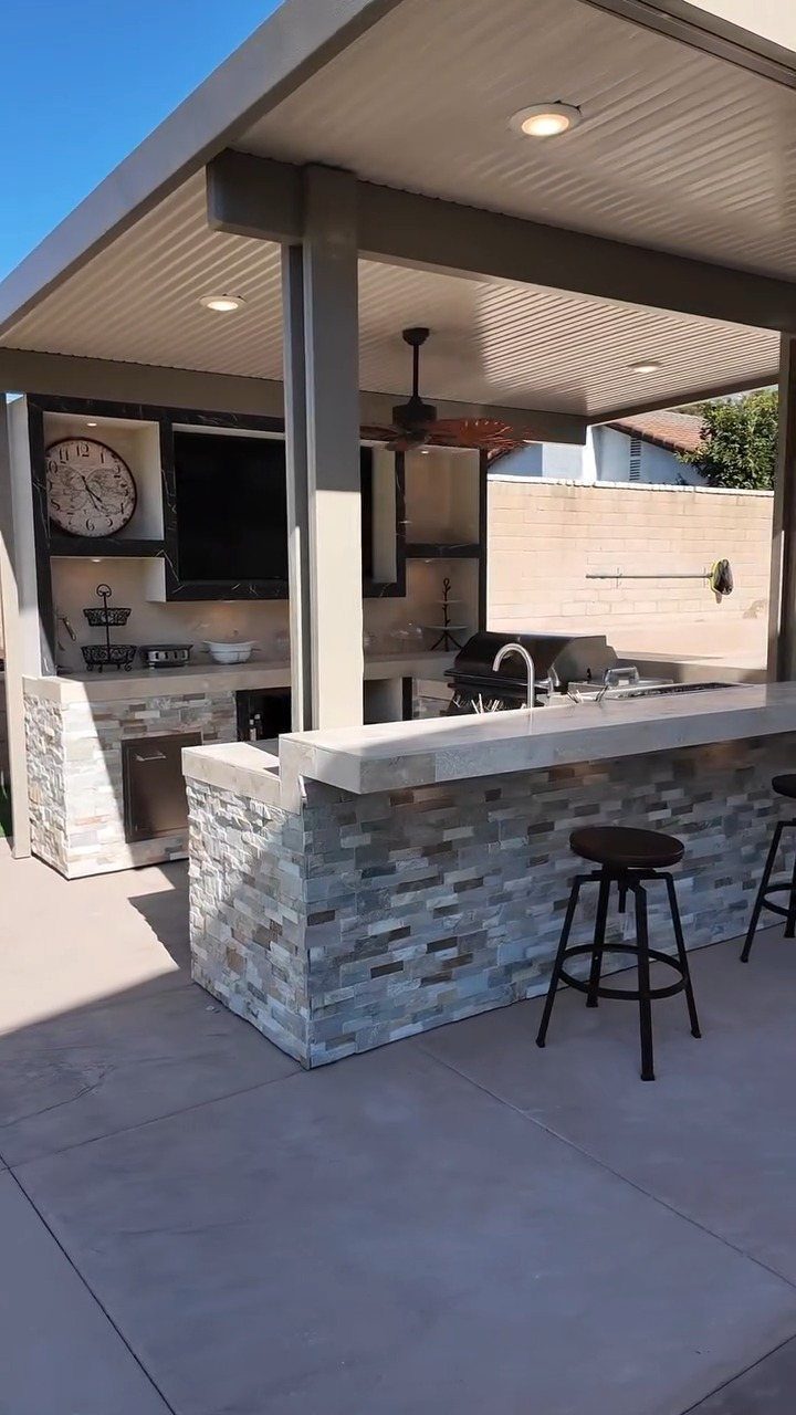A stone-clad U-shaped outdoor kitchen with a built-in grill and bar seating under a patio cover.