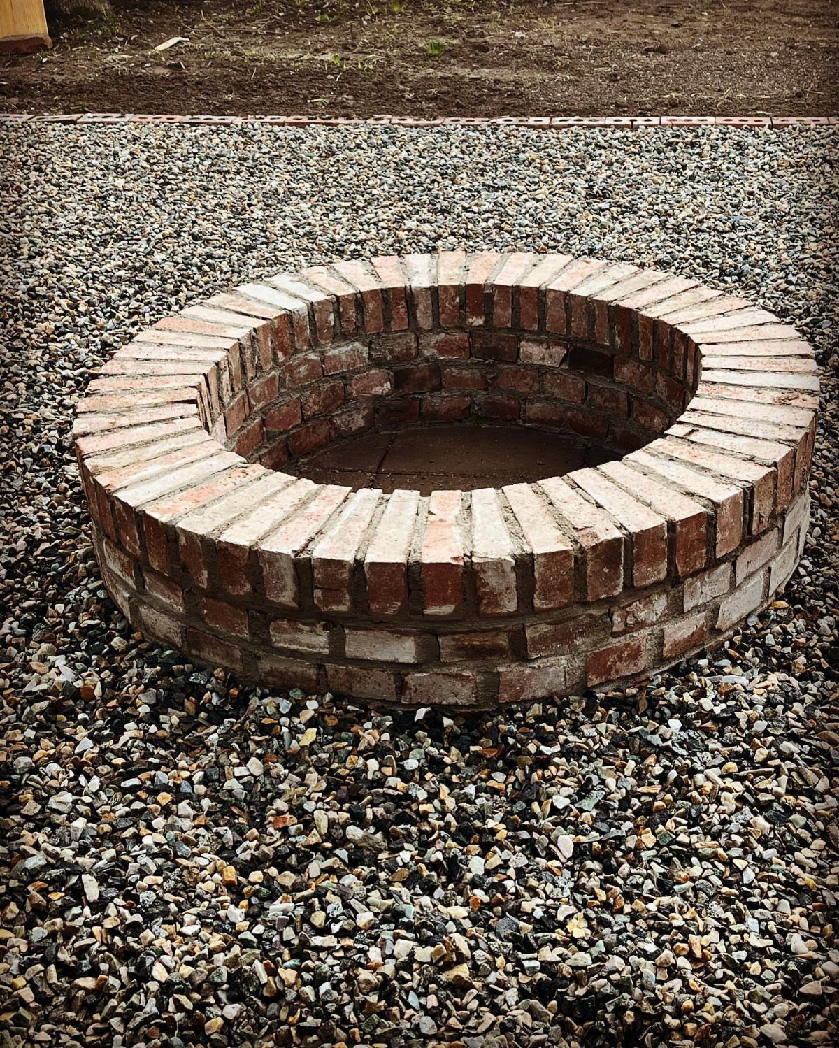 A circular brick fire pit sits embedded in a bed of small grey gravel stones.